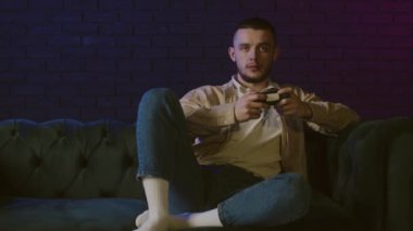 Konsol oynayan genç bir adam. Erkek siber sporu, evde oyun oynamak. O şimdi yeni bir oyun bölümü başlattı. Genç bir adam kanepeye uzanıp neon ışıklarla video oyunları oynuyor. Yüksek kalite