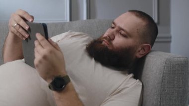 Beyaz sakallı genç adam oturma odasındaki tabletten içeriği izliyor. Evde yalnız başına zaman geçir. Yetişkin adam kanepede oturuyor ve tablet kullanıyor. Rahat erkek kanepeye uzanıp film izliyor. Yüksek
