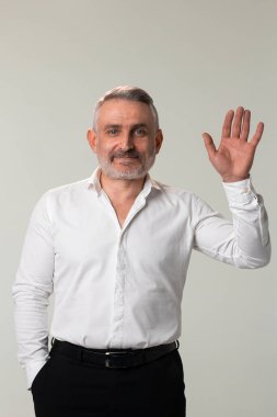A man dressed in formal wear, consisting of a white dress shirt and black suit trousers, is waving his hand in a gesture while showcasing his beard and arm