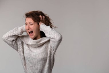 A woman in a sweater is loudly screaming while clutching her hair in distress, her jaw clenched and sleeves rolled up in a gesture of frustration