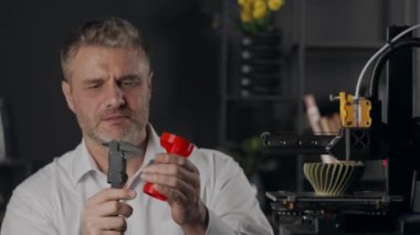 Orta yaşlı bir grafik tasarımcısı dijital kalibreler kullanır ve kırmızı plastik huninin parametrelerini ölçer. Otomatik özerk süreçte çalışan ve haki rengi üreten 3D yazıcı makinesi