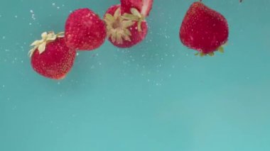 Turkuaz bir arka planda bütün ve yarı yarıya kırmızı çilek parçaları dökülüp suya batar, yüzeye çıkar ve kabarcıklar oluşturur. Yavaş çekim