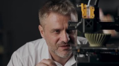 Orta yaşlı bir grafik tasarımcısı 3D yazıcının çalışma sürecini gözlemler. Bir makine haki renkli geometrik şekil yapar. Modern üretim teknolojisi kavramı. Otomatik olarak üç boyutlu