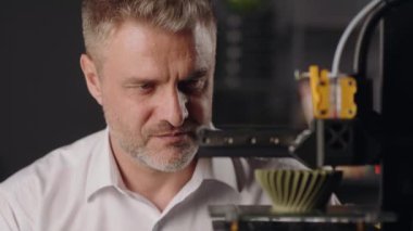 Orta yaşlı bir grafik tasarımcısı 3D yazıcının çalışma sürecini gözlemler. Bir makine haki renkli geometrik şekil yapar. Modern üretim teknolojisi kavramı. Otomatik olarak üç boyutlu