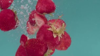 Turkuaz bir arka planda bütün ve yarı yarıya kırmızı çilek parçaları düşer ve su sıçrar, kabarcıklar ve damlalar oluşturur, suya batar ve suyun altında yüzer. Yavaş