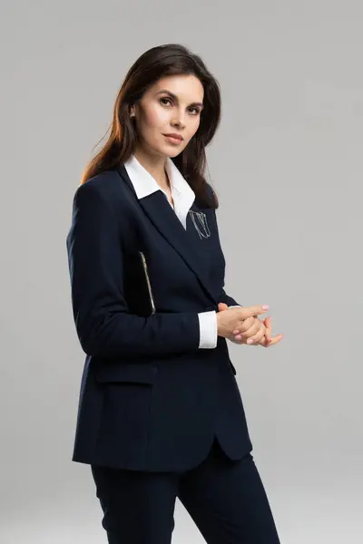 Takım elbiseli bir kadın elleri katlı, ceketli bir gömlek giyiyor. Saçları düzgünce yapılmış ve profesyonel görünüyor..