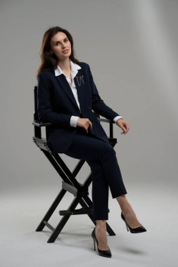 Elektrikli mavi ceketli kadın yönetmen koltuğunda oturuyor, bacağını çaprazlıyor ve rahatlamak için dizini sandalyede dinlendiriyor..