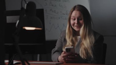 Genç sarışın bir kadın iş yerinde masada oturuyor, işten dinleniyor, akıllı telefon kullanıyor, ekrana yakından bakıyor, okuyor, gülümsüyor ve hayret verici bir sürprizle karşılaşıyor.