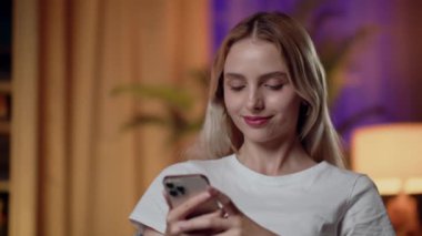 Genç bir kadın sıcak bir şekilde oturma odasında gülümsüyor, dijital iletişim için akıllı telefonu kullanıyor.