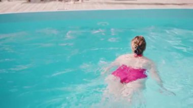 Güneşli bir günde mavi havuzda yüzen pembe mayo giymiş bir kadın. Bir tatil köyünde rahatlık ve rahatlık..