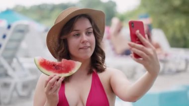 Havuz kenarında karpuzla selfie çeken şık şapkalı bir kadın eğlenceli bir yaz tatilini simgeler.