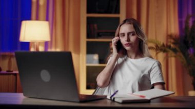 Genç bir kadın ev ofisinde birden fazla iş yapıyor, telefonla konuşuyor, dizüstü bilgisayarda çalışıyor, notlar yazıyor, gece geç saatlerde..