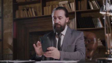 Takım elbiseli sakallı bir işadamı kitaplıklarla çevrili geleneksel bir ofiste akıllı telefon kullanıyor.