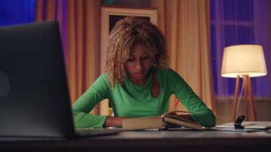 Odaklanmış yeşil bluzlu bir kadın kitap ve notlarla çalışıyor rahat bir ev ortamında, akşam vakti..