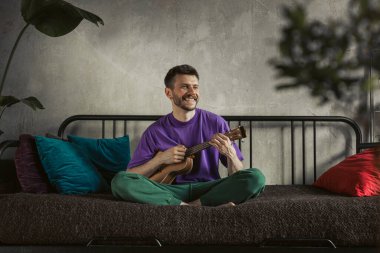 Mor gömlekli bir adam, parlak yastıklar ve dekorlarla çevrili şık bir kanepede ukulele çalar.