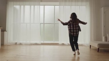 Parlak, güneşli, ahşap zeminli ve büyük pencereli bir odada dans eden gündelik kıyafetli bir kadın..