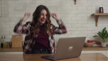 Mutfak ortamında dizüstü bilgisayar kullanan neşeli bir kadın başarıyı veya başarıyı kutluyor..