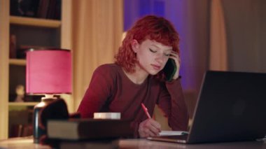 Kızıl saçlı bir kadın dizüstü bilgisayar ve telefonda birden fazla iş yapıyor, notlar alıyor ve rahat bir odada evden çalışıyor.