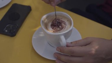Yakın plan çekimde, biri köpüklü bir kapuçinoyu yanında akıllı telefonu olan sarı bir masada beyaz bir bardakta karıştırıyor.