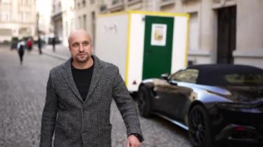 Arka planda modern bir spor arabasıyla Avrupa 'da bir kaldırım taşı caddesinde yürüyen gri ceketli bir adam..