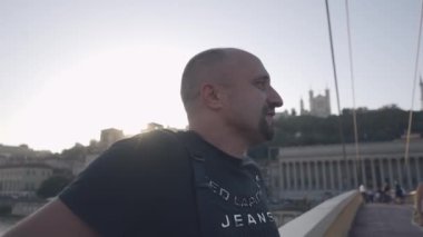 Sakallı kel adam köprü korkuluklarında dikiliyor, şehir manzarası var, kot gömlek giyerek günbatımının tadını çıkarıyor..