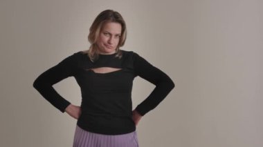 Stüdyoda elleri kalçasında, siyah bir bluz ve mor bir etek giyen bir kadın..