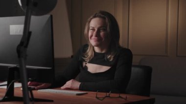 Enerjik bir kadın modern ofis ortamında masasında gülüyor ve çalışıyor, ekranla meşgul oluyor..