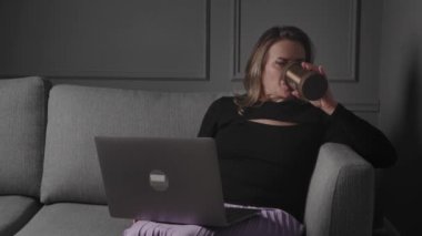 Bir kadın, gri bir kanepede sıcak bir içecek içerken dizüstü bilgisayarında çalışıyor ve evinde rahat bir ortam yaratıyor..