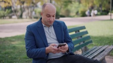 Orta yaşlı bir adam parkta bir bankta oturuyor, akıllı telefonuna dalmış, doğal ortamında keyfine bakıyor.