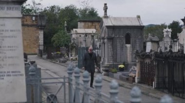 Güneş gözlüklü bir adam karanlık gökyüzünün altındaki tarihi mezarlıkta gotik mezarların ve mezar taşlarının arasında yürüyor.