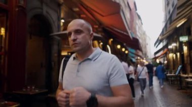 Kel bir adam kalabalık bir Avrupa caddesinde yürüyor kafeler ve restoranlarla dolu, hayat dolu şehir atmosferinin tadını çıkarıyor.