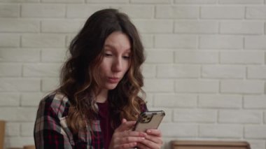 Ekose gömlekli, uzun saçlı genç bir kadın akıllı telefon kullanarak, tuğla bir duvarın önünde durarak hayrete düşmüş görünüyor.