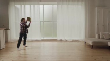 Bir kadın geniş bir minimalist oturma odasında duruyor, büyük pencerelerin yakınındaki bir defter ya da cihaza atıfta bulunuyor..