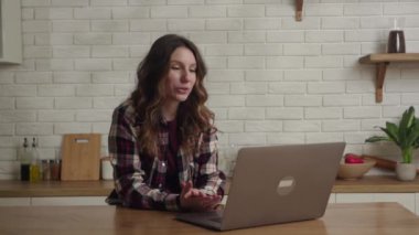 Modern bir mutfakta, bir kadın uzaktan çalışma ve video çağrısı için dizüstü bilgisayar kullanır, telekomünikasyon eğilimlerini gösterir