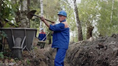 Mavi üniformalı ve güvenlik teçhizatlı çalışkan bir inşaat işçisi şantiyede kürekle çukur kazıyor.