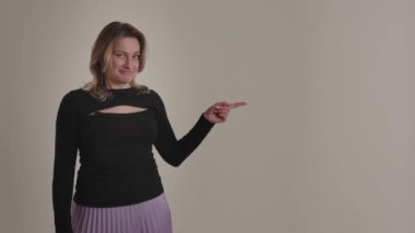Genç, neşeli bir Amerikalı ya da Avrupalı sarışın kadın gülümser, avuçlarını yana uzatır, işaret parmağı yukarıda ve bej arka planda boş bir alan gösterir. Bir bayan teşvikçi neşe saçar.