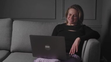 Genç, gülümseyen Amerikalı ya da Avrupalı bir kadın blogcu evdeki kanepede oturur, el sallayarak selamlama ve işaret eder, sosyal ağda video çağrısı için bir dizüstü bilgisayar kullanır,