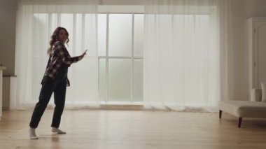 Büyük pencereleri ve perdeleri olan, parlak, ferah bir oturma odasında dans eden ve eğlenen bir kadın..