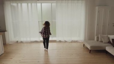 Modern bir oturma odasında, ahşap zemini ve geniş penceresi olan bir kadın tek başına neşeyle dans ediyor..