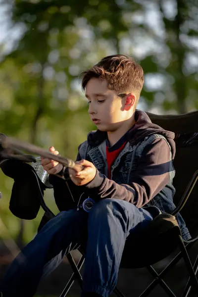 Genç bir çocuk elinde olta tutarken rahat bir şekilde bir sandalyede oturuyor, balık tutmak ve dışarıda eğlenmek için sabırsızlanıyor.