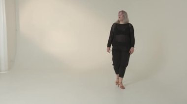 Kendine güvenen bir kadın minimalist bir stüdyo ortamında zarif ve güzel bir şekilde yürüyor, modern moda eğilimlerini sergiliyor.