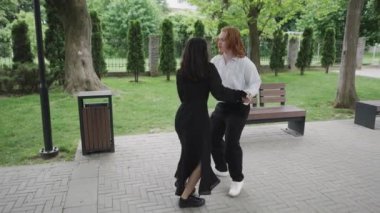 Canlı bir parkta, iki kadın kaldırımda arkadaşlığı neşeyle kucaklarken, bir çift yakınlarda dans ediyor, büyüleyici atmosferi güçlendiriyor.