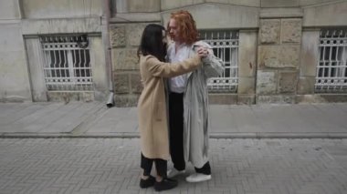 Canlı bir şehirde, trençkotlu kendine güvenen bir kadın sıcak bir kucaklaşmayla arkadaşını selamlar. Neşeli zamanları, şehir yaşamının ortasındaki dostluğu vurguluyor.