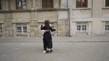 Yoğun bir kaldırımda, iki kadın neşeyle dans ediyor, dikkat çekiyorlar. Çiftler sevgi paylaşıp sıcak bir hava yaratırken bir erkek de katılıyor.