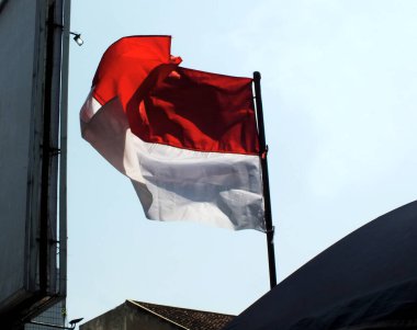 Indonesia national flag waving in the wind between city building and public facility with clear blue sky background. Nation patriotism. The Hari Kemerdekaan Indonesia clipart