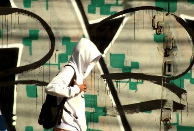 Kapüşonlu ve sırtında sırt çantası olan biri duvar yazısıyla duvarın önünden geçiyor. Graffiti çoğunlukla yeşil ve siyah, bazı beyaz harflerle. Manzara biraz gergin ve kentsel..