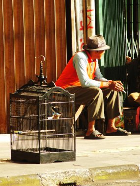 Bandung City, Batı Java, Endonezya, 25 Ocak 2025. Adam kuş kafesinin yanındaki bankta oturuyor. Adam turuncu bir yelek giyiyor.