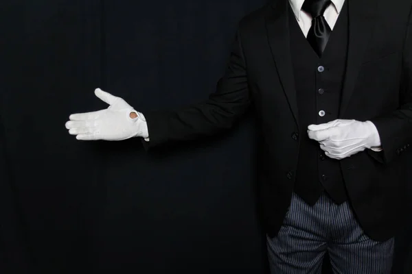 Portrait of Butler or Servant in Dark Suit and White Gloves with Welcoming Gesture on Black Background. Service Industry. Professional Hospitality and Courtesy. Formal White Glove Service.
