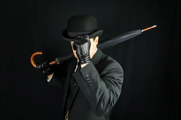 Gentleman in Dark Suit Tipping Bowler Hat on Black Background. Concept of Classic and Eccentric British Gentleman Stereotype. Film Noir Secret Agent Spy
