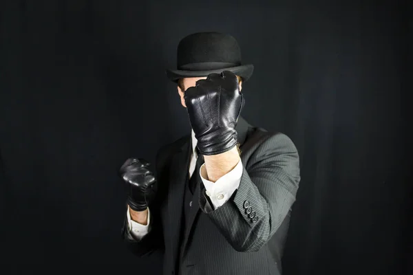 Gentleman in Dark Suit and Leather Gloves with Fists Raised in Fighting Pose on Black Background. Concept of Classic and Eccentric British Gentleman Stereotype. Film Noir Secret Agent Action Hero